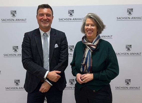 Zwei Personen stehen lächelnd vor einer Pressewand mit dem Schriftzug „Sachsen-Anhalt“ und dem Landeswappen. Ein Mann im dunklen Anzug mit Krawatte und eine Frau mit grünem Oberteil und Schal posieren nebeneinander bei einem offiziellen Termin.